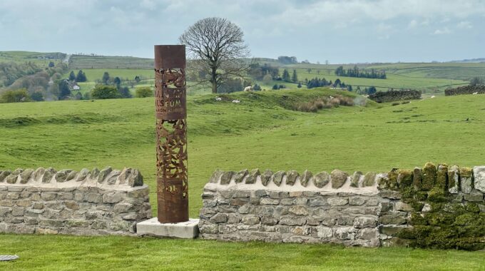 Grass, a wall and a sculptural beacon with a field and lone tree beyond.