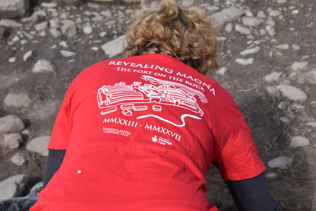 Volunteer excavator in the trench with their back to the camera showing their Magna Ts-shirt