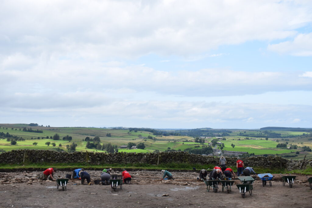 Landscape view of the archaeological trench at Milecastle 46 with the hills of Cumbria in the background. Wheelbarrows and excvators are in the foreground. 