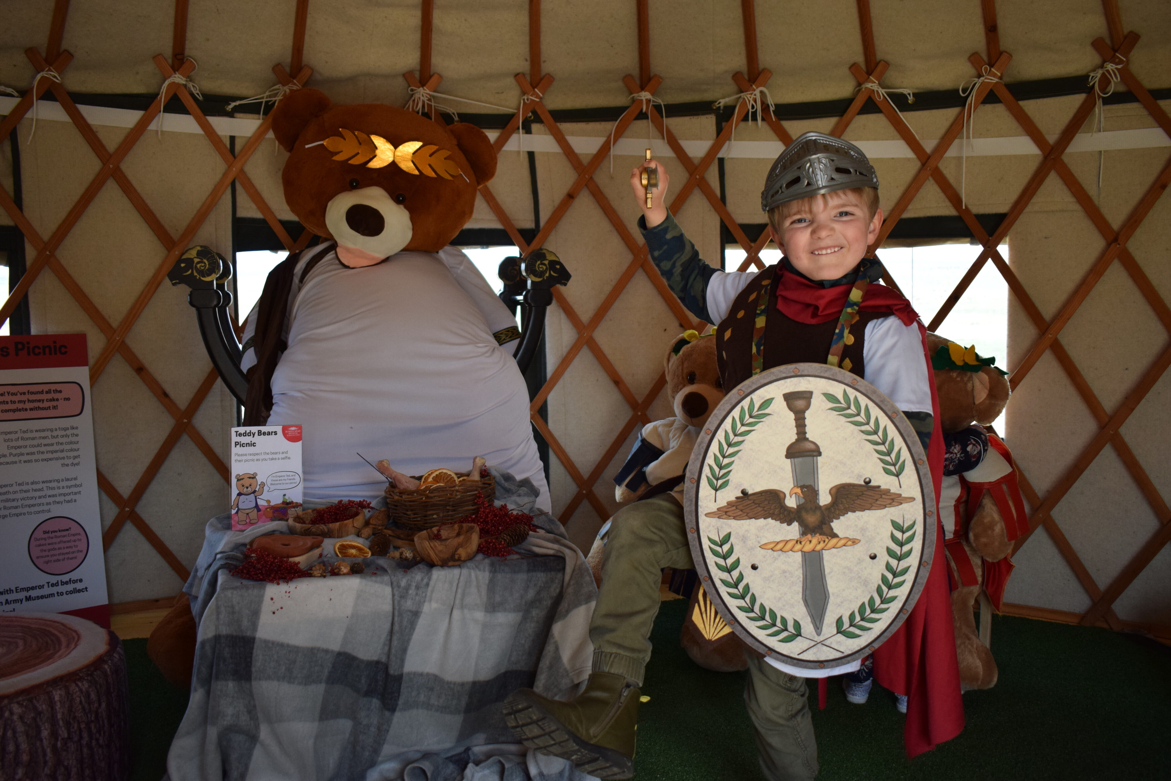 A young boy dressed in a Roman soldier costume, including a helmet, cape, and shield, stands next to a large teddy bear dressed as a Roman emperor seated on a throne.
