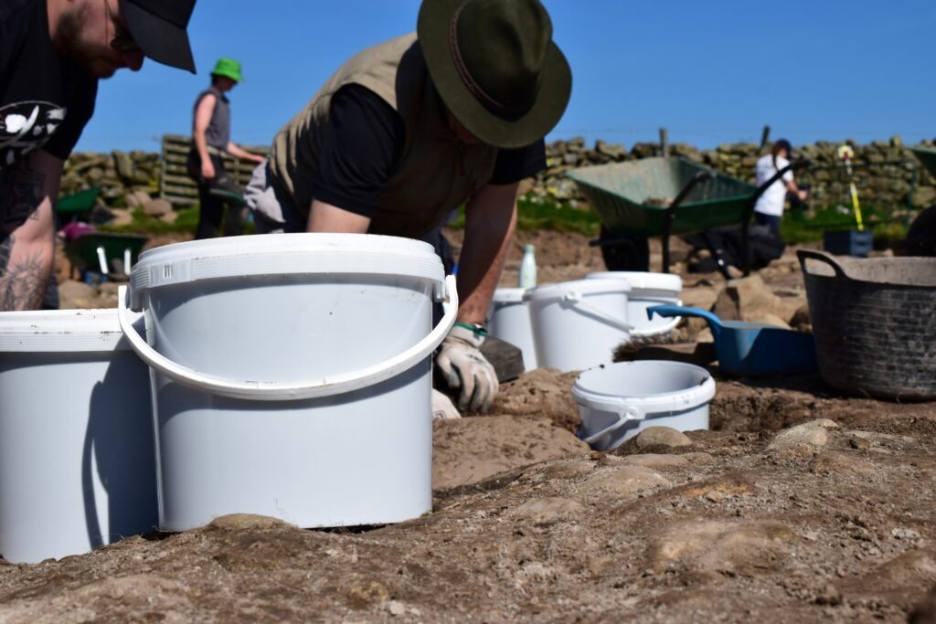 White soil sample buckets with archeaological excavations in background.