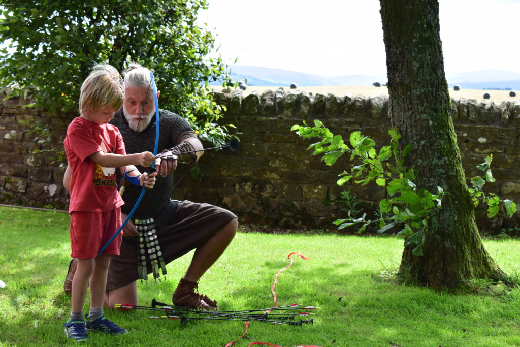 Man with silver hair and a beard dressed as Roman archer shows a young boy how to hold a bow and arrow.