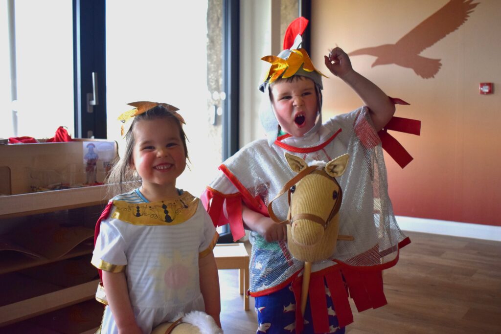 An image of two young children dressed up as a Roman Soldier and Roman emperor holding hobby horses smiling at the camera.