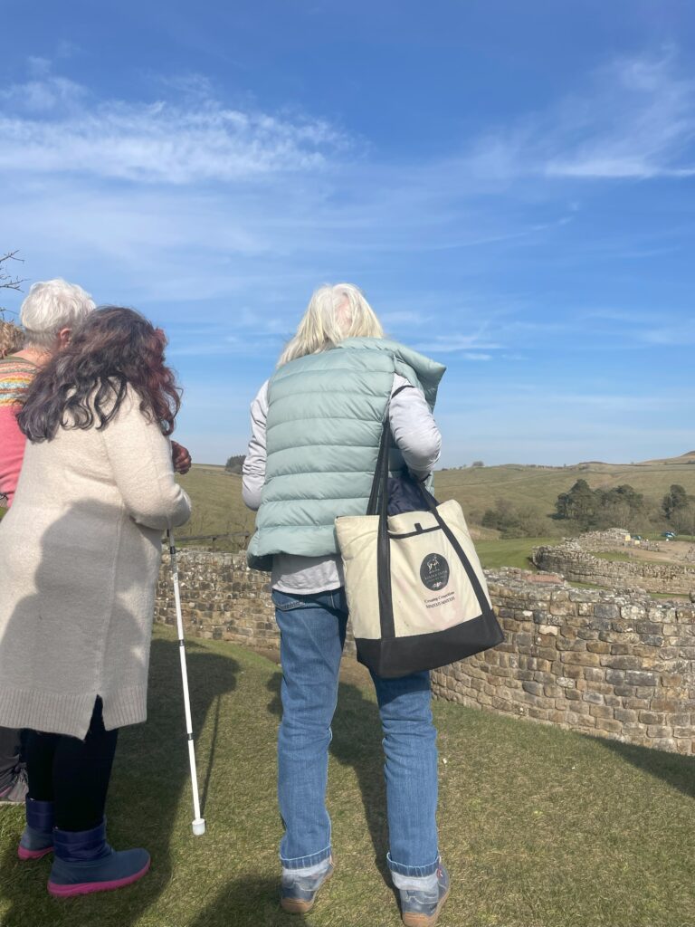 Image of two people standing with their back to the camera facing ancient roman fort ruins. The person on the right has long brown hair, is wearing a beige tunic jumper and holding a white cane. The person on the left has short, white hair, a green gilet, and blue jeans.