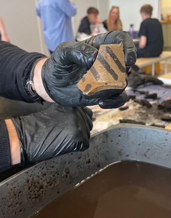 Image of black gloves holding a sherd of brown Roman pottery with black stripes running diagonally across it.