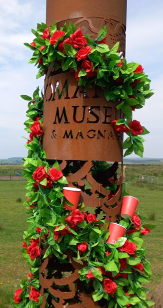Image of a metal beacon with the word 'Roman Army Museum & Magna Fort' carved into it. The beacon is covered in roses and several red cups are poking out of the roses.