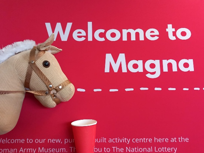 Image of a hobby horse to the left, pretending to take a drink from a red cup. This is on a red background with the words 'Welcome to Magna' written.