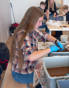 Image of a white girl with long brown hair sitting side on. She is wearing blue gloves and is washing pottery in a bowl.