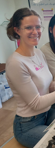 Image of a white girl with brown hair, a pink top, jeans, and glasses. She is smiling at the camera.