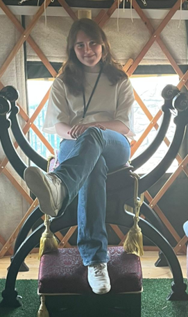 Image of a white girl with shoulder length brown hair sitting on an ornamental chair. She is facing the camera with her legs crossed, smiling.