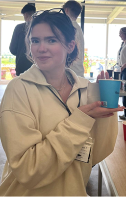 Image of a white girl with brown hair wearing a cream quarter zip jumper. She is holding a blue cup and smiling at the camera.