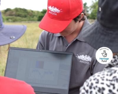 A group of people with their backs to the camera are looking at a laptop screen in a grassy field. The person holding the laptop has a grey polo shirt, red cap, and short brown hair.