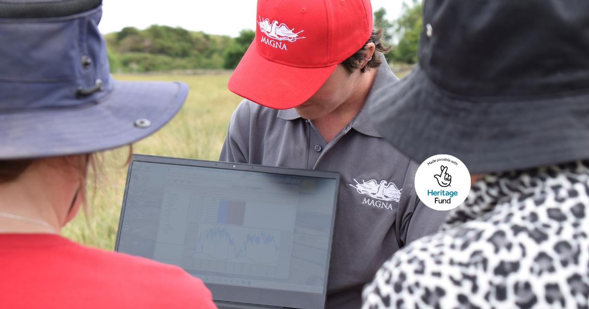 A group of people with their backs to the camera are looking at a laptop screen in a grassy field. The person holding the laptop has a grey polo shirt, red cap, and short brown hair.