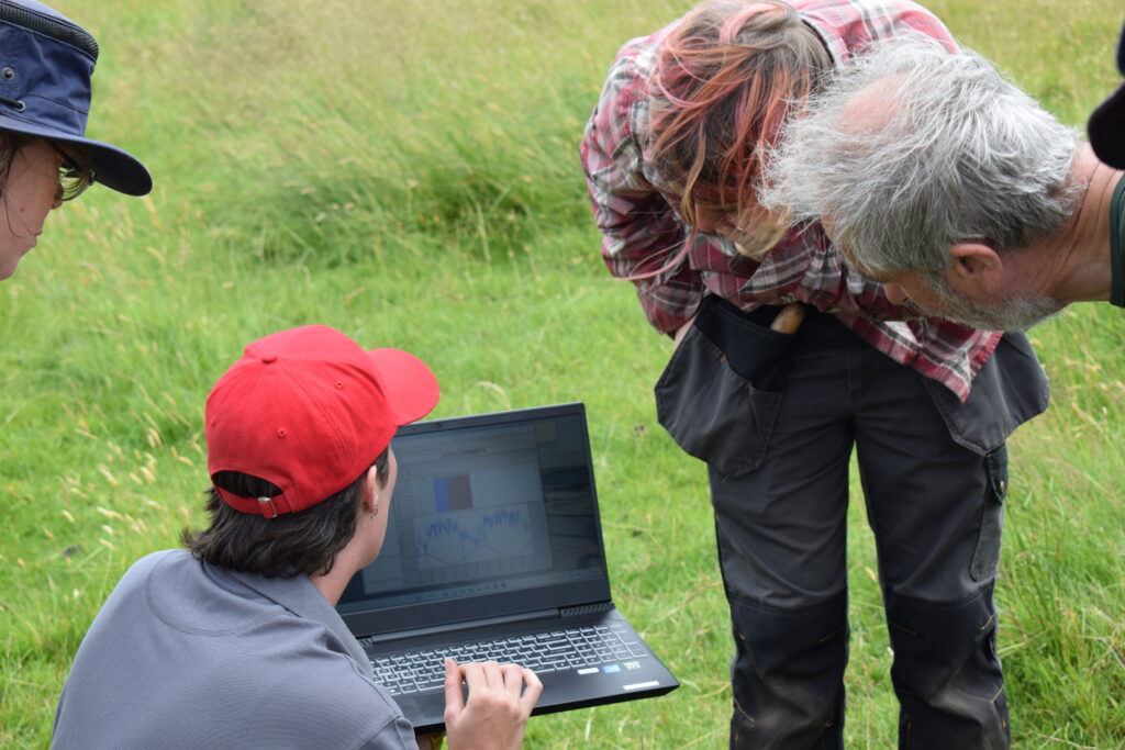 A group of people with their backs to the camera are looking at a laptop screen in a grassy field. The person holding the laptop has a grey polo shirt, red cap, and short brown hair.
