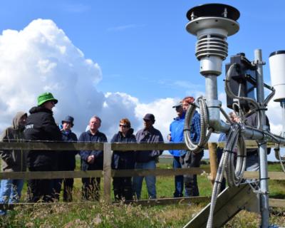 In the foreground is a weather monitoring station, with a number of cables, panels, and scientific instruments used to measure changes in the weather. A group of people stand behind it, leaning on a fence listening to a talk about the station.