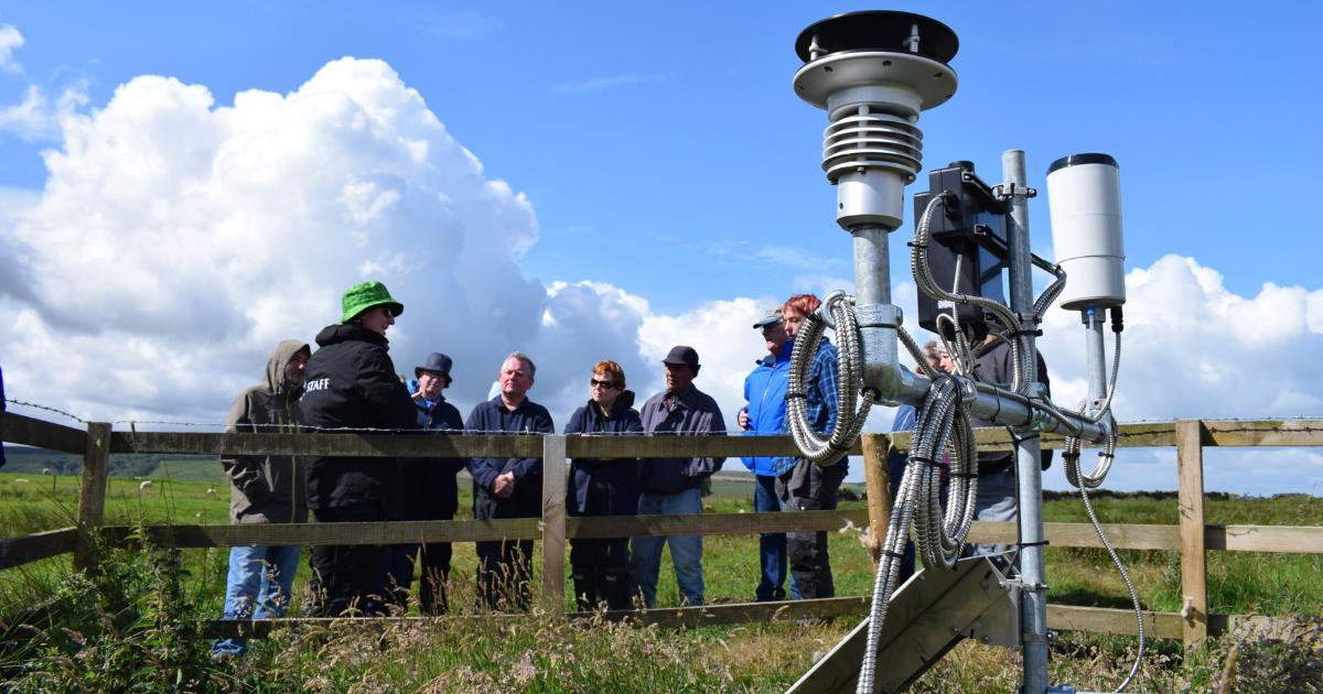 In the foreground is a weather monitoring station, with a number of cables, panels, and scientific instruments used to measure changes in the weather. A group of people stand behind it, leaning on a fence listening to a talk about the station.