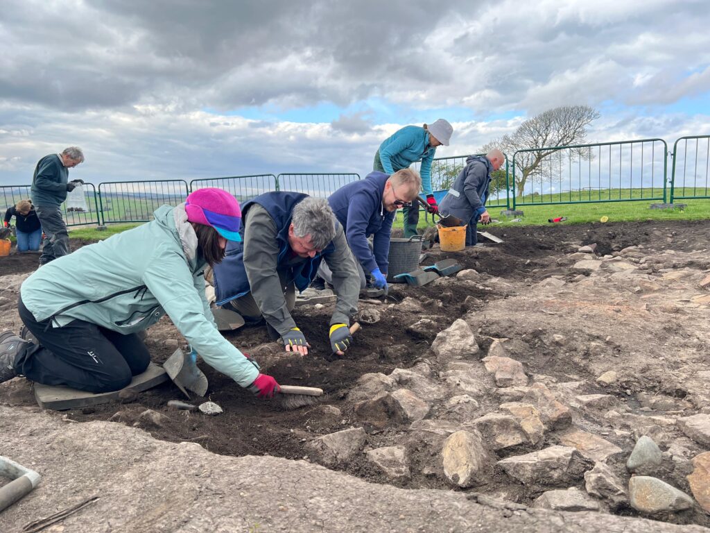Volunteers carefully excavating a stone-lined archaeological trench using brushes and trowels, with open countryside visible in the background.