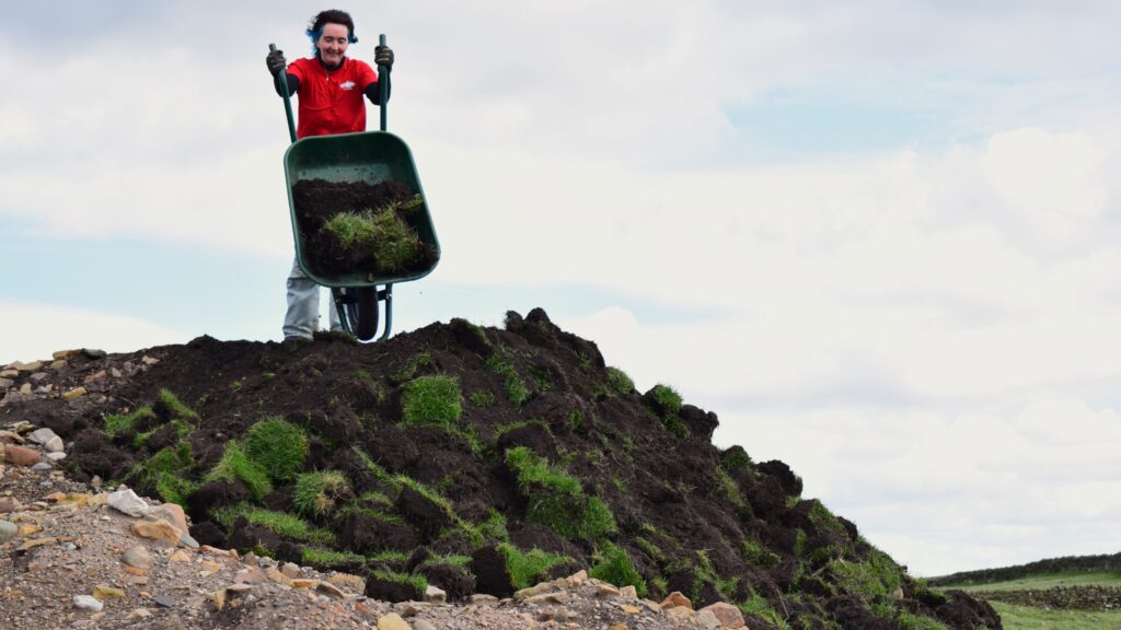 A woman with blue hair and a red t-shirt is tipping a wheelbarrow full of turf and soil onto a spoil heap. The angle of the picture is from the ground up. There is cloudy sky in the background. 