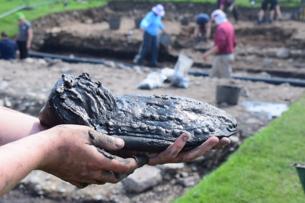 Hands holding a Roman leather shoe showing the hobnails that remain on the sole. In the background are archaeological excavators working at Magna Roman Fort.