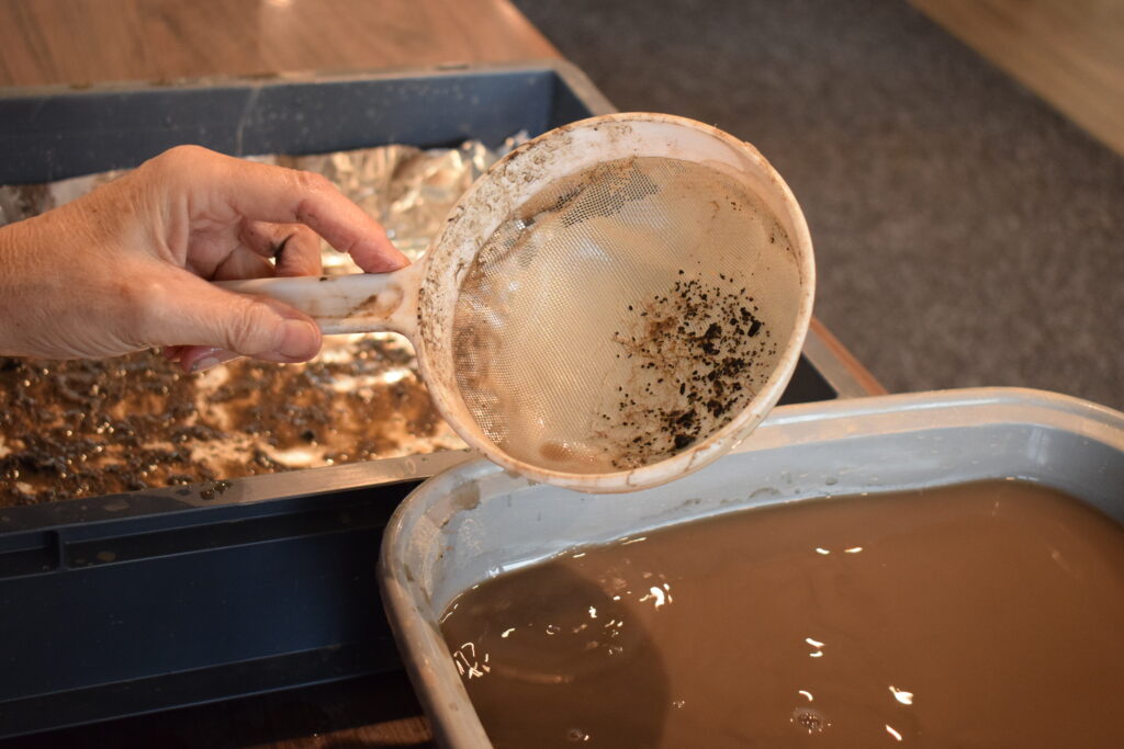 A hand holds a fine mesh sieve over a tray of muddy water during wet sieving of excavated soil, used to recover small archaeological finds or environmental samples.