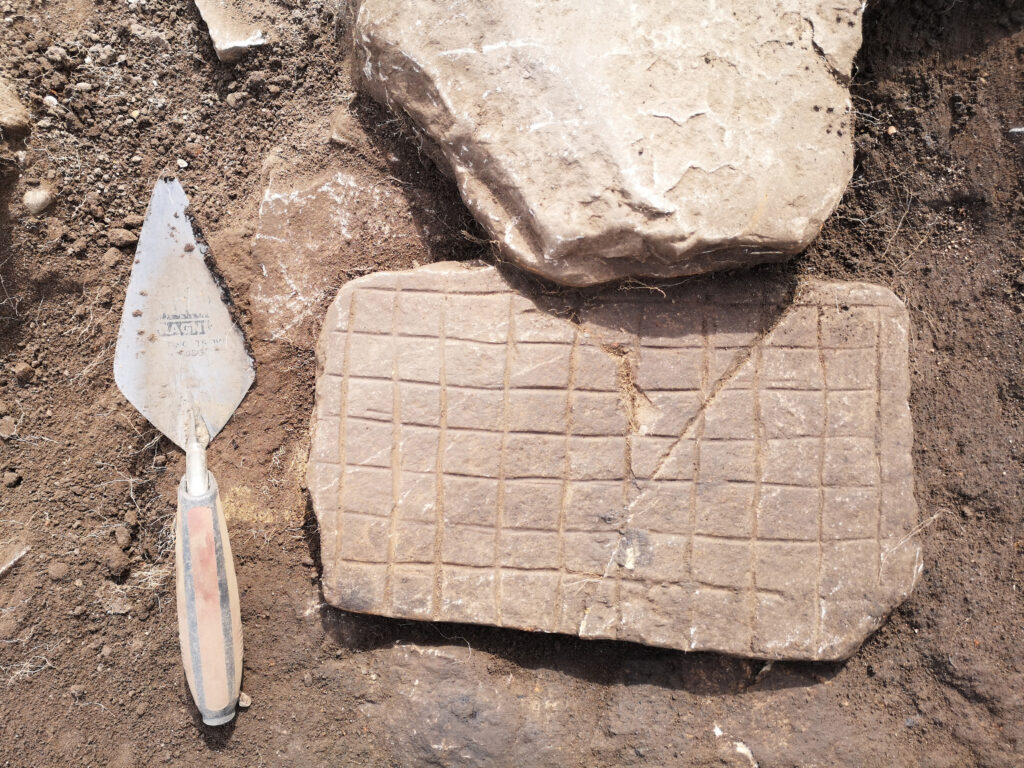An archaeological find showing a flat stone slab with a grid of carved lines forming rows and columns of squares, resembling a gaming board, photographed next to a trowel at an excavation site.