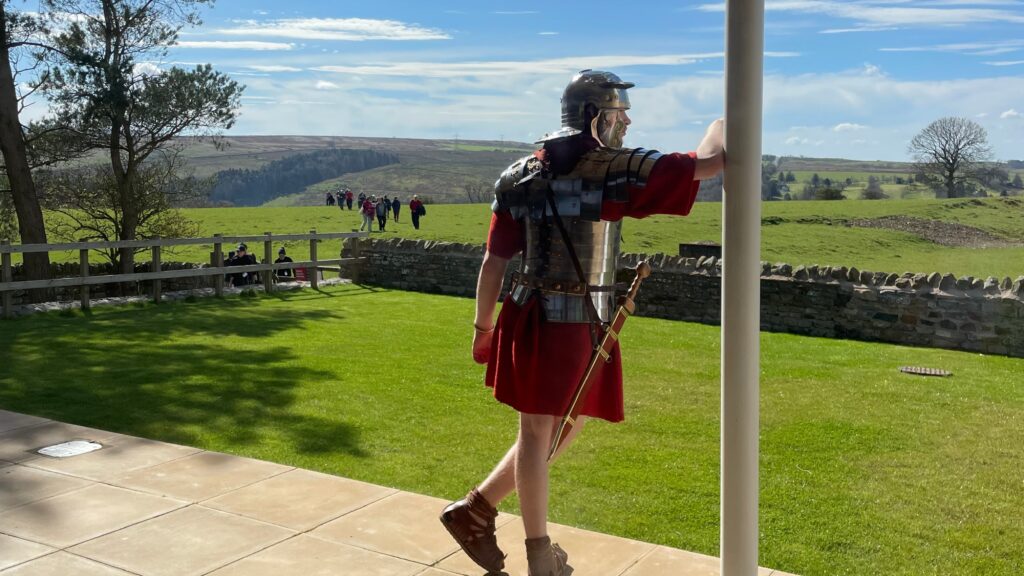 A Roman legionary reenactor in full lorica segmentata armour, red tunic and gladius stands at the Roman Army Museum, overlooking the Hadrian's Wall landscape at Magna Roman Fort, Northumberland.