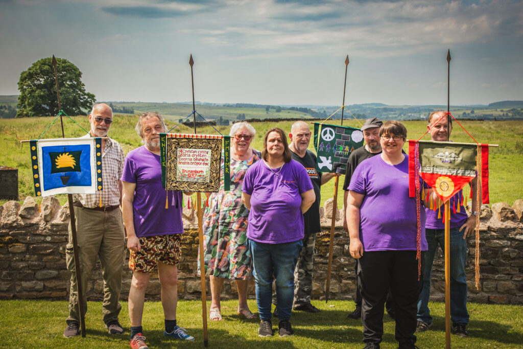 A group poses outdoors holding handmade decorative banners on spear-topped poles, including one referencing the Battle of Brunanburh, with rolling green countryside behind them.