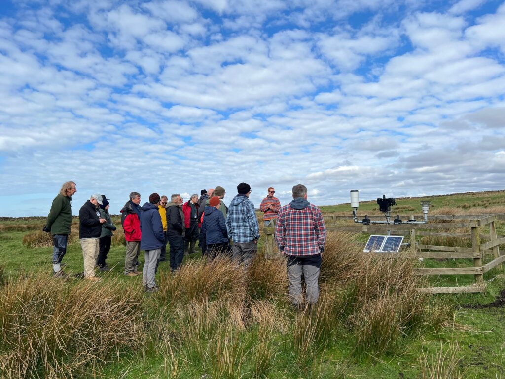 A group of people gathered around environmental monitoring equipment on open moorland under a cloudy blue sky.