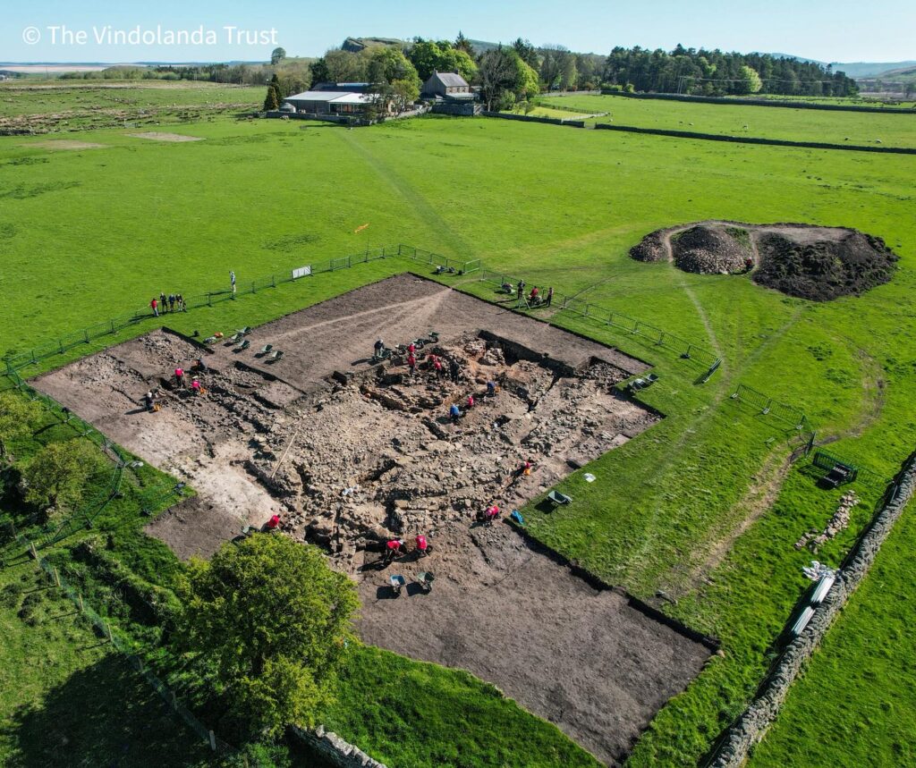 Aerial view of volunteers excavating the Magna Roman Fort trench on a sunny day, with extensive stone remains visible across the site and green Northumberland countryside beyond.