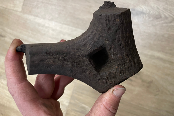 A hand holding a dark brown fragment of worked antler with a square hole cut through it, likely a roman gaming dice.