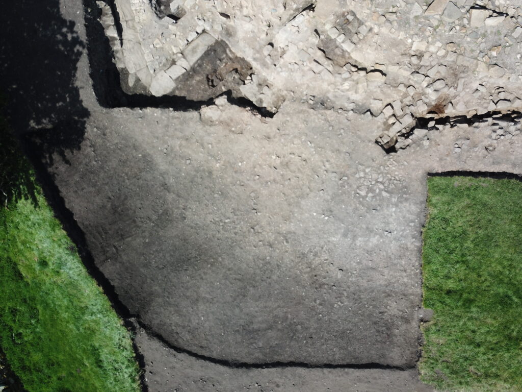 Aerial close-up of an archaeological excavation trench showing a flat compacted surface with scattered small stones, bordered by green grass and partially collapsed dry-stone walling. There is a soil colour differentiation curving along the line of a roman defensive ditch.