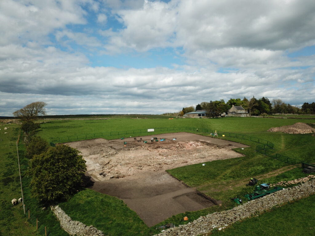 Aerial view of a large rectangular archaeological excavation site in a green rural landscape, with exposed stone foundations, equipment, and dry-stone boundary walls visible.