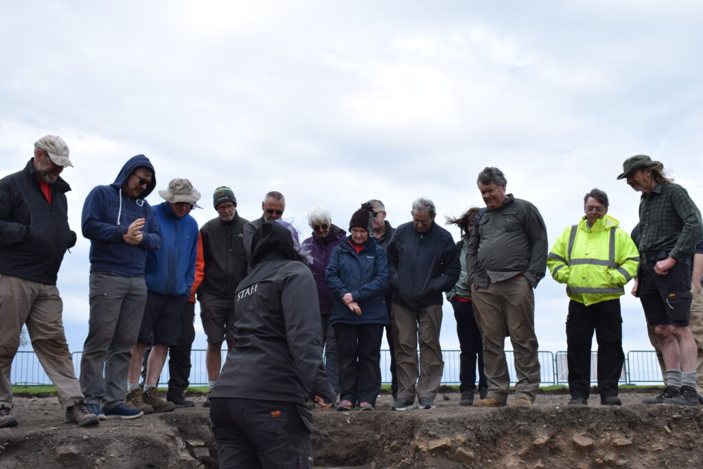 A staff member explains findings to a group of visitors gathered around an archaeological excavation trench on an overcast day.