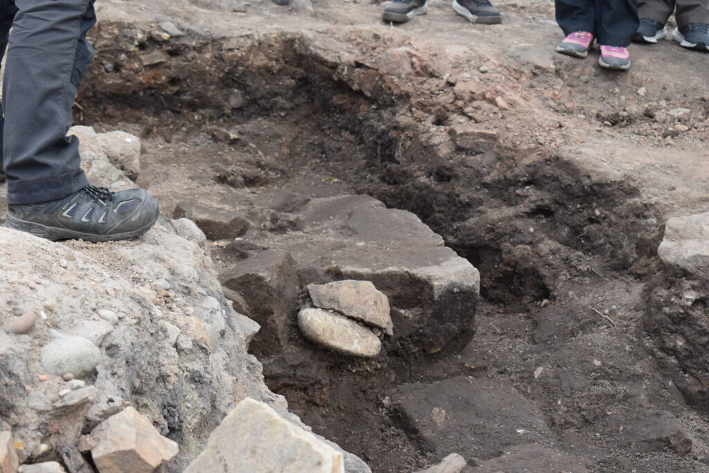 Close-up of exposed stone features in an archaeological excavation trench, with archaeologists feet visible at the edges.