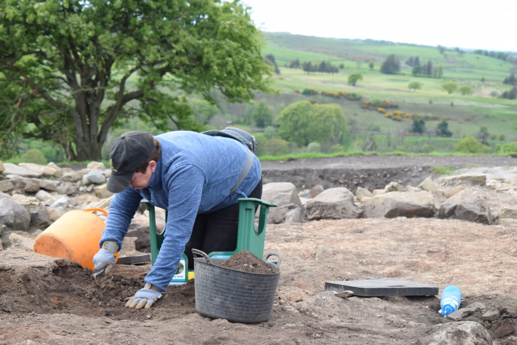 An archaeological volunteer in a blue top and cap kneels to excavate a trench by hand, with a trowel and spoil buckets nearby, surrounded by exposed stonework against a backdrop of rolling green countryside.