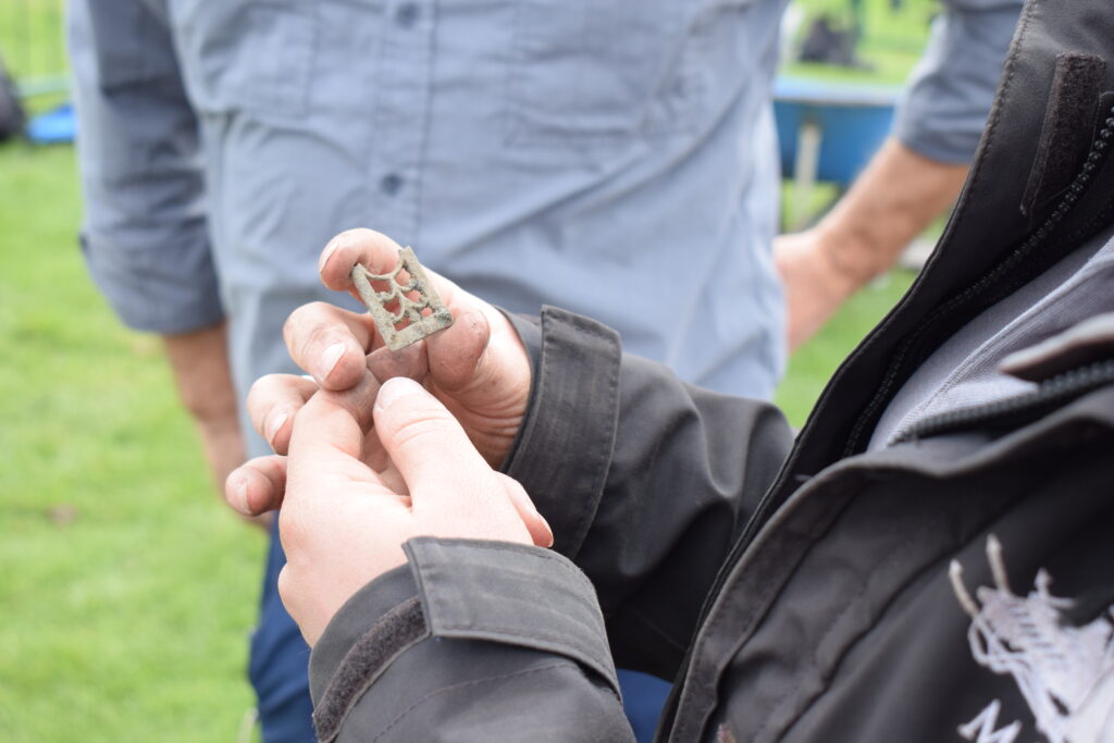 Two people standing on grass examining a small corroded metal artefact with openwork decoration, possibly an archaeological find or metal detecting discovery, held up for closer inspection.
