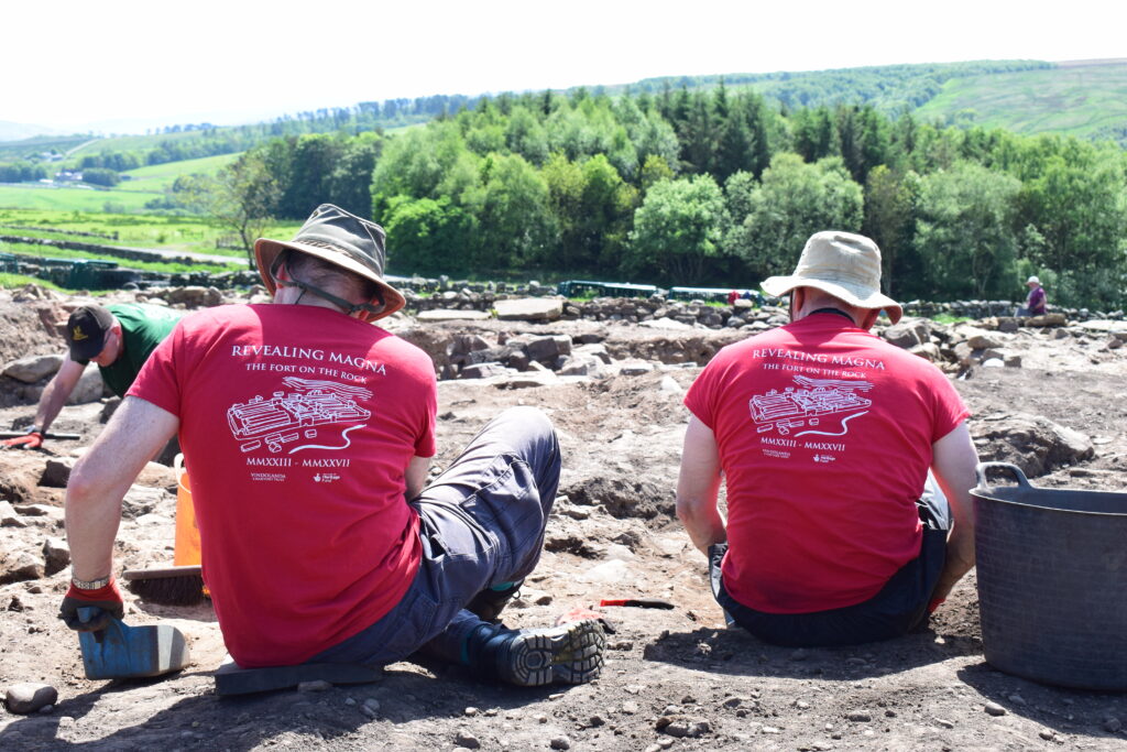16:28Claude responded: Two volunteers in "Revealing Magna – The Fort on the Rock" t-shirts excavating a Roman archaeological site, with stone ruins, rolling hills and woodland in the…Two volunteers in "Revealing Magna – The Fort on the Rock" t-shirts excavating a Roman archaeological site, with stone ruins, rolling hills and woodland in the background.