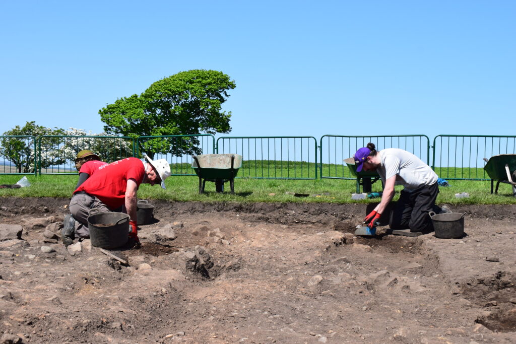Two archaeologists excavating a dig site on a sunny day, kneeling in a rectangular trench with trowels and buckets, green fields and wheelbarrows visible behind a metal fence.