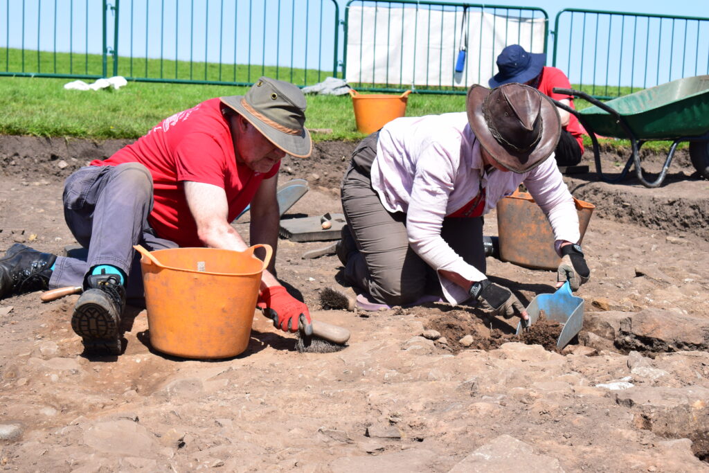 Two archaeologists in wide-brimmed hats kneeling in the Magna excavation trenches, carefully excavating a stone surface with trowels and brushes.