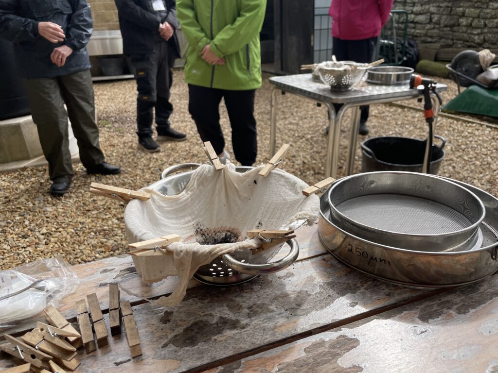 A muslin cloth secured with wooden clothes pegs over a colander, used for straining, alongside metal sieves labelled "250mm", on an outdoor worktable during a practical demonstration of floatation in archaeological processing.