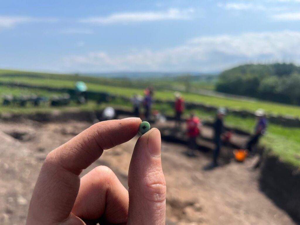 A hand holds up a small green bead at an archaeological dig site, with excavation trenches and team members visible in the background against a rolling green landscape.
