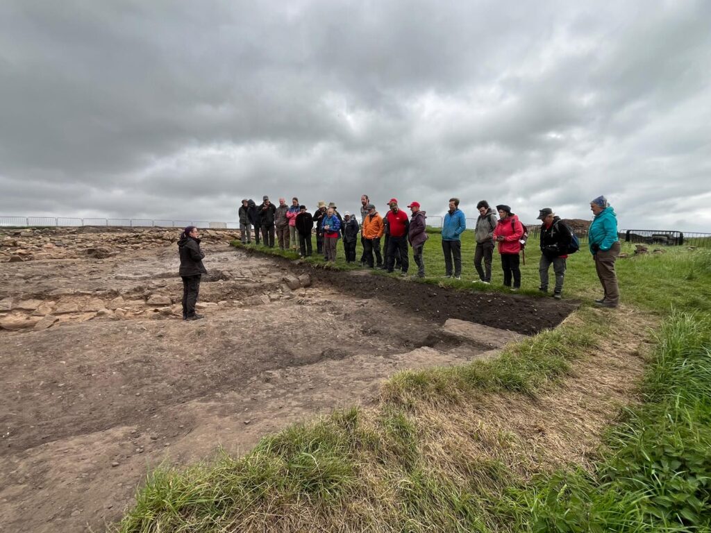 An archaeologist addresses a group of visitors standing at the edge of an open excavation trench, with stone ruins and an overcast sky in the background.