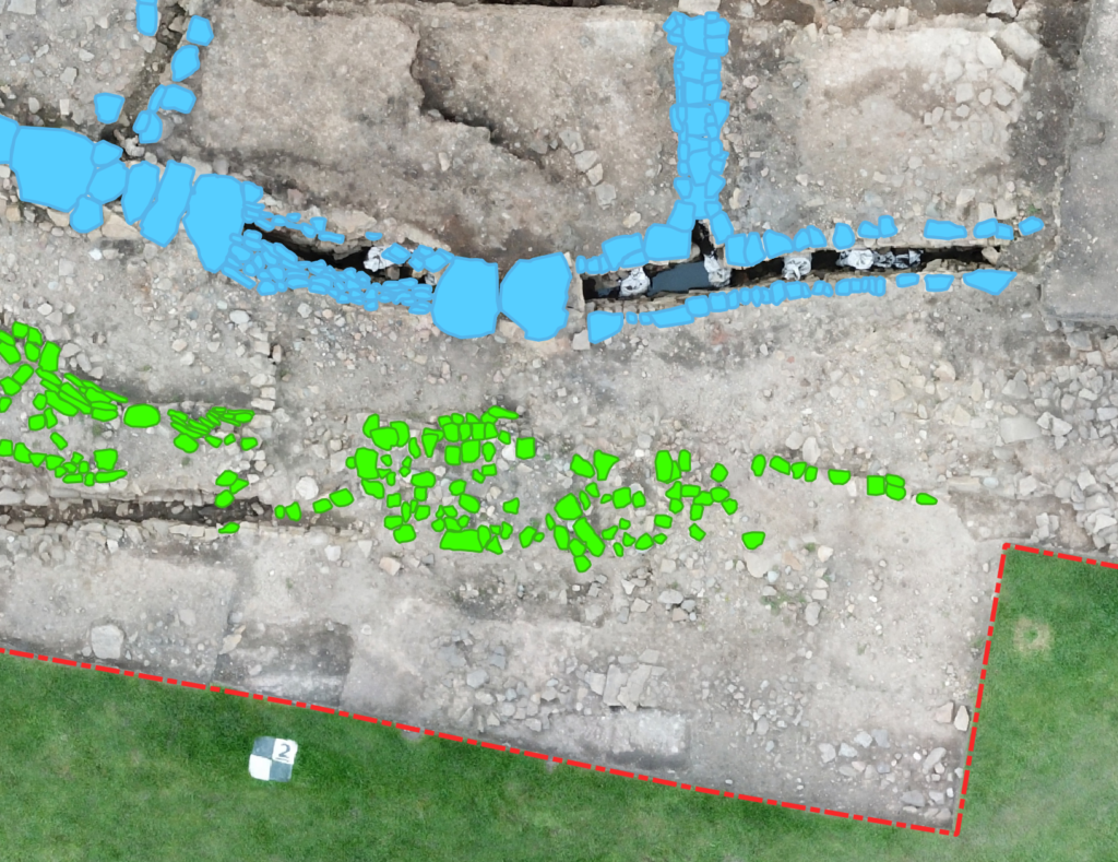 An aerial view of an archaeological excavation site with QGIS vector overlays: blue polygons marking larger stones along a wall feature, green polygons highlighting smaller stones along a second feature, and a red dashed rectangle outlining a trench boundary. A scale marker labelled number 2 is visible in the lower left area.