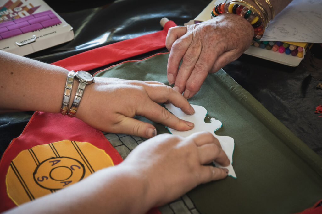 Close-up of hands positioning a felt cut-out shape onto a green fabric banner during its construction, with red and yellow fabric pieces visible nearby.