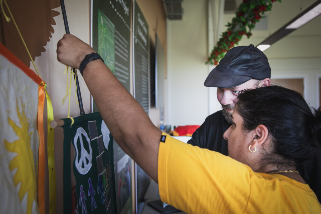 Two people fit a hand-painted banner featuring a peace symbol and figures onto a display pole, standing beside informational panels indoors.