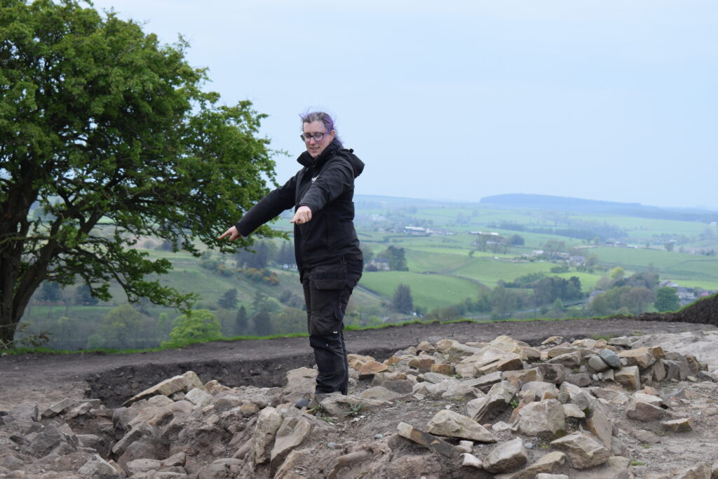 An archaeologist points out features among exposed stone remains at an excavation site, with a sweeping green valley landscape visible behind them.