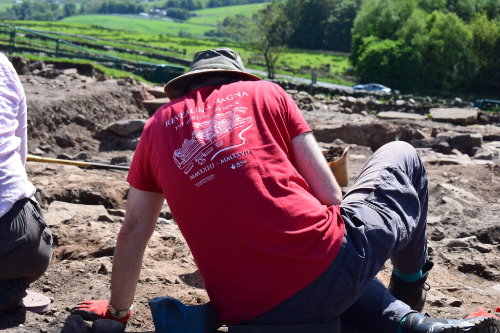 Person sitting in an archaeological trench with their back to the camera. They are wearing a red t-shirt which reads 'Revealing Magna - the Fort on the Rock'