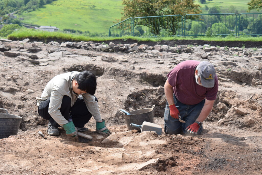 Two archaeological volunteers in the foreground crouching on the ground with trowels and brushes on an archaeological site. Rolling green hills and trees are in the background. 