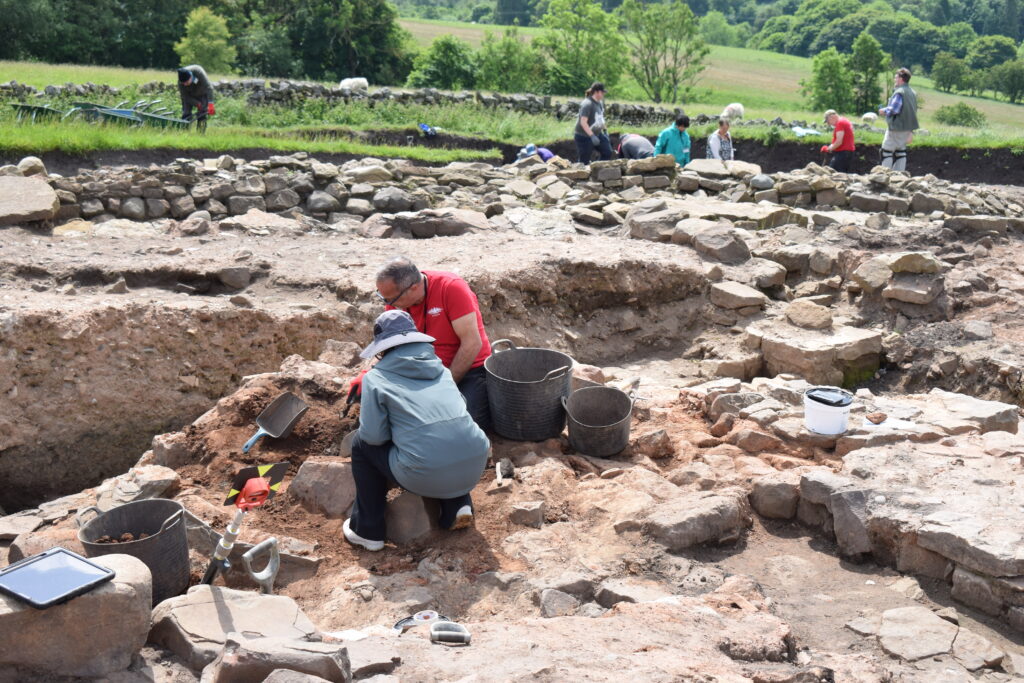 Two archaeologists work closely together in a trench surrounded by exposed stone walls, with a wider team excavating across the sun-lit site and green countryside beyond.