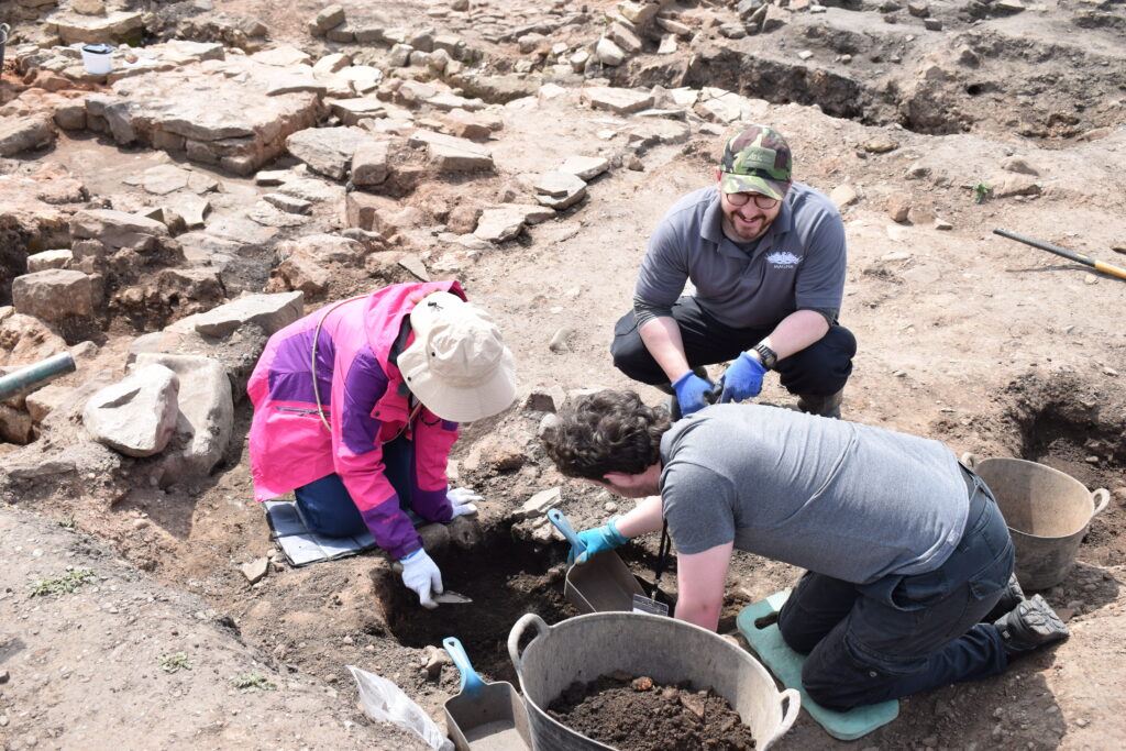 Two archaeology volunteers and the Magna archaeological assistant excavating a pit feature on an archaeological site. They are using scoops and buckets. 